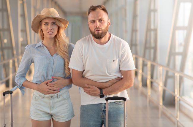 Egy fehér pólós férfi és egy kék inges nő áll a reptér csarnokában, hasukat fogva, fájdalmas arckifejezéssel.