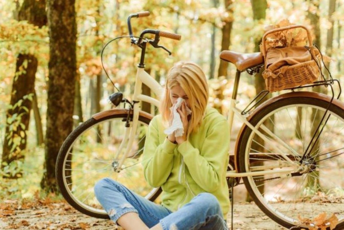 Egy nő zöld pulóverben ül a földön az erdőben, és egy fehér zsebkendőbe fújja az orrát. Mögötte egy bicikli.